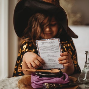 Puede incluir: Un ni&ntilde;o vestido de bruja sostiene una tarjeta que dice "Stir Up Your Power" con instrucciones para hacer una poci&oacute;n m&aacute;gica. La tarjeta enumera ingredientes como "una pizca de polvo de sapo", "una pizca de valor de bruja", "1 cucharadita de humor m&aacute;gico", "1 cucharada de madera ligera", "caldero irrompible", "peque&ntilde;o frasco de vidrio" y "una cuchara de madera".