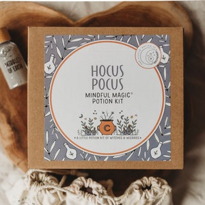 Puede incluir: Una caja marr&oacute;n con una etiqueta blanca que dice "Hocus Pocus Mindful Magic Potion Kit" con una caldera de dibujos animados y estrellas. La caja est&aacute; sobre una superficie de madera con una peque&ntilde;a botella de purpurina.