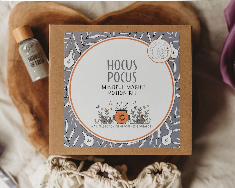 Puede incluir: Una caja marr&oacute;n con una etiqueta blanca que dice "Hocus Pocus Mindful Magic Potion Kit" con una caldera de dibujos animados y estrellas. La caja est&aacute; sobre una superficie de madera con una peque&ntilde;a botella de purpurina.