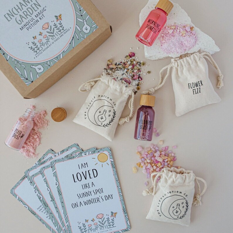 Puede incluir: Un kit de pociones m&aacute;gicas conscientes con una caja marr&oacute;n, cinco frascos peque&ntilde;os, cinco bolsitas peque&ntilde;as y cinco tarjetas de afirmaci&oacute;n. Los frascos contienen l&iacute;quidos rosas, rojos y transparentes. Las bolsitas contienen cristales rosas, blancos y transparentes. Las tarjetas tienen fondos verdes y blancos con afirmaciones impresas en ellas. La caja tiene el texto "Enchanted Garden Mindful Magic Potion Kit" impreso en ella.