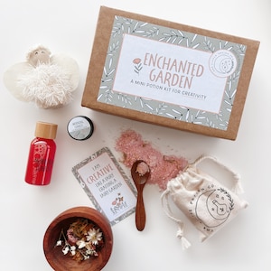 May include: A brown box with the text "ENCHANTED GARDEN A MINI POTION KIT FOR CREATIVITY" on the front. The box contains a small white bag with the text "THE LITTLE POTION CO." on it, a small wooden bowl with dried flowers, a small spoon, a small bag of pink bath salts, a small bottle of red liquid, a small card with the text "I AM CREATIVE LIKE A FAIRY CRAFTING A FAIRY GARDEN", and a small white angel figurine.