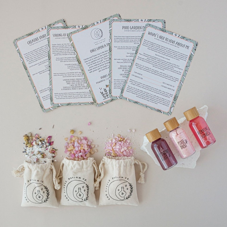 Puede incluir: Tres peque&ntilde;as bolsas de muselina blancas llenas de sales de ba&ntilde;o rosas, cada una con un logotipo en blanco y negro que dice "The Little Potion Co.". Tres peque&ntilde;as botellas de vidrio con tapas de corcho llenas de l&iacute;quidos rosas y rojos. Las botellas est&aacute;n etiquetadas como "Pixie Dust", "Morning Sauna" y "Dancing Fernande". Cinco tarjetas con fondos verdes y texto blanco, con t&iacute;tulos como "Hada creativa", "Fuerte de", "&Eacute;rase una vez una luna", "Jard&iacute;n de hadas" y "Lo que no te gusta de m&iacute;".
