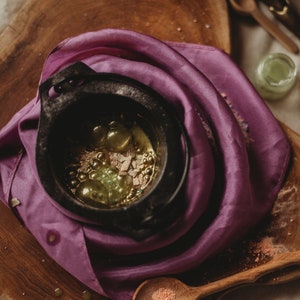 Puede incluir: Un caldero negro con un pa&ntilde;o morado drapeado sobre &eacute;l. El caldero est&aacute; lleno de una mezcla de l&iacute;quido verde y dorado y peque&ntilde;os trozos de material blanco.
