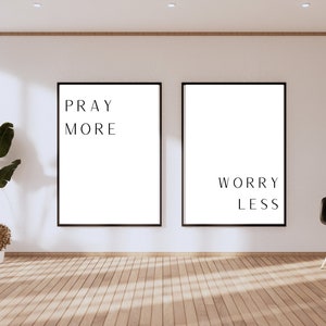 May include: Two black framed prints with white backgrounds and black text. The left print says "Pray More" and the right print says "Worry Less". The prints are hanging on a white wall in a room with wooden floors.