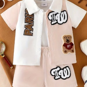 Op de afbeelding: Een tweedelige baby-outfit in roze en wit. Het shirt heeft een witte kraag en een knoopsluiting aan de voorkant. Het shirt heeft een bruine "PARIS"-patch aan de linkerkant en een witte "TW"-patch aan de rechterkant. De broek heeft een witte "TW"-patch aan de voorkant en een bruine teddybeer-patch met het nummer "87" aan de achterkant.