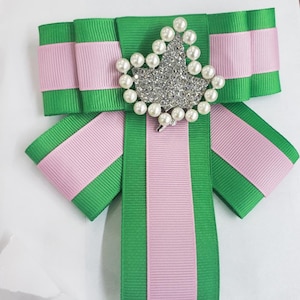 Puede incluir: Un lazo de rayas verdes y rosas con un broche de hoja de perlas y diamantes de imitación.