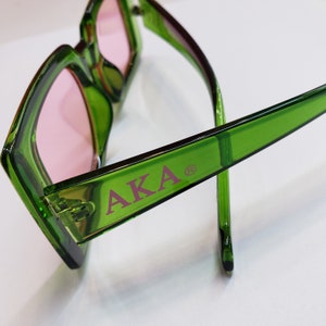 May include: Green square-shaped sunglasses with pink lenses. The sunglasses have the word "AKA" printed in pink on the side.