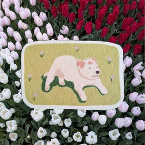 May include: A rectangular rug with rounded corners featuring a light yellow background with small pink flowers and a central illustration of a light pink dog lying down on a patch of dark green grass with a simple black eye and nose.