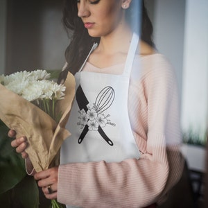 May include: A white apron with a black and white design of a knife and whisk crossed over each other with a floral design. The apron is worn by a person holding a bouquet of white flowers.