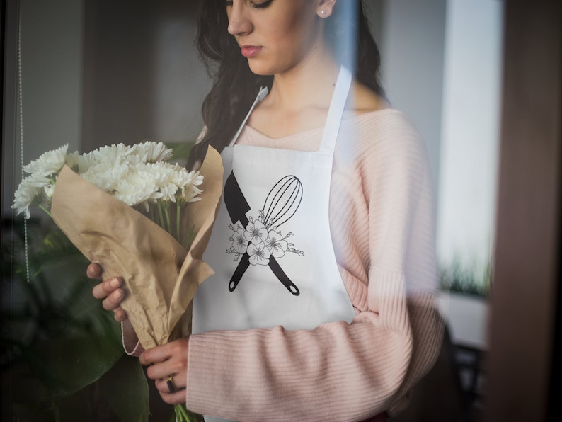 May include: A white apron with a black and white design of a knife and whisk crossed over each other with a floral design. The apron is worn by a person holding a bouquet of white flowers.