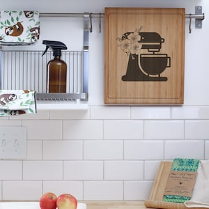 May include: A kitchen countertop with a white tiled backsplash. There are two red apples on a white rectangular plate. A wooden cutting board with a green and white patterned beeswax wrap set is on the counter. The cutting board has the text "Beeswax Wrap Set" printed on it. A wooden cutting board with a floral design and a silhouette of a stand mixer is hanging on the wall.