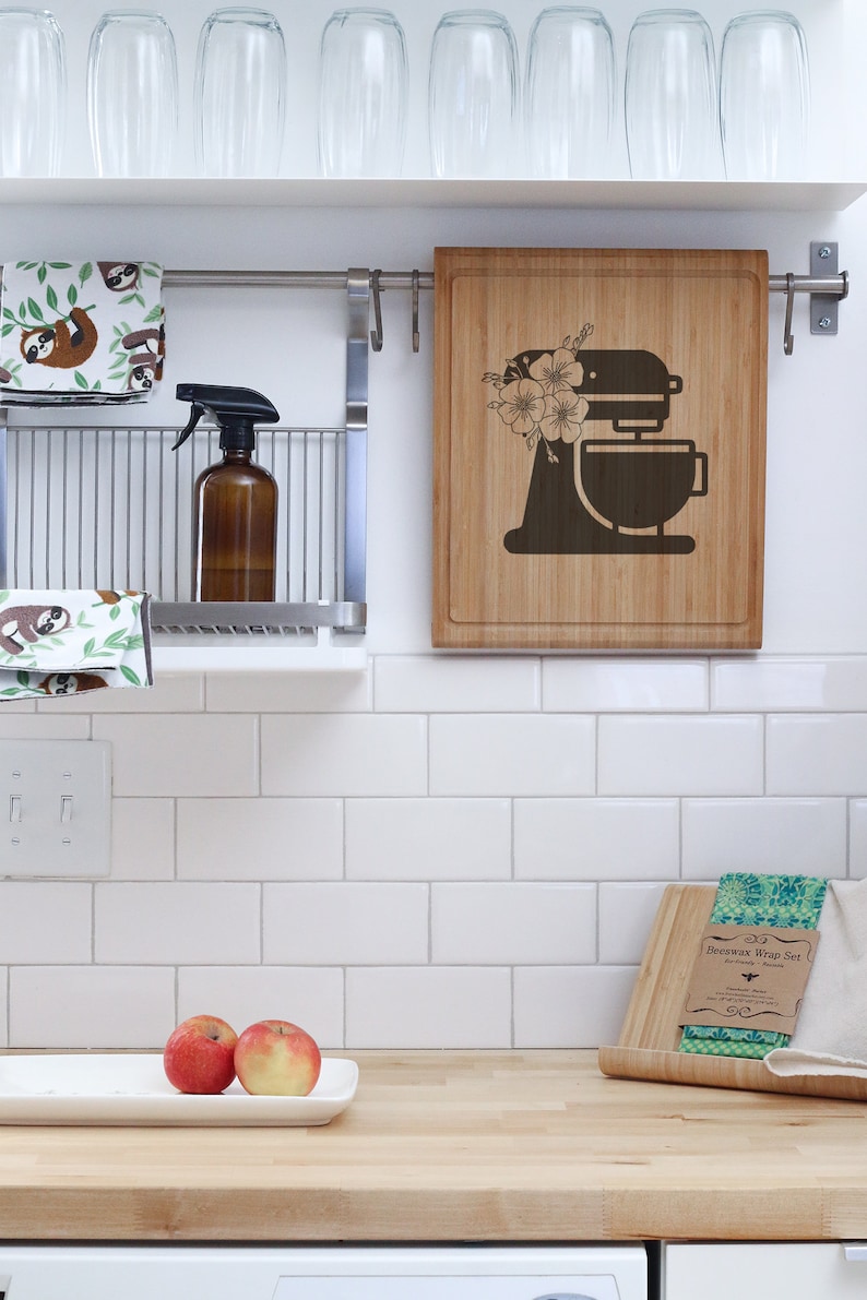 May include: A kitchen countertop with a white tiled backsplash. There are two red apples on a white rectangular plate. A wooden cutting board with a green and white patterned beeswax wrap set is on the counter. The cutting board has the text "Beeswax Wrap Set" printed on it. A wooden cutting board with a floral design and a silhouette of a stand mixer is hanging on the wall.