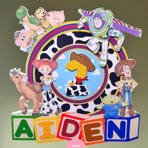 K&ouml;nnte beinhalten: Bunte Tortenaufsatz mit Toy Story-Figuren, darunter Woody, Buzz Lightyear und Jessie. Das Design beinhaltet den Namen "AIDEN" in Blockbuchstaben und die Zahl eins. Eine lebendige Dekoration f&uuml;r eine Geburtstagstorte.