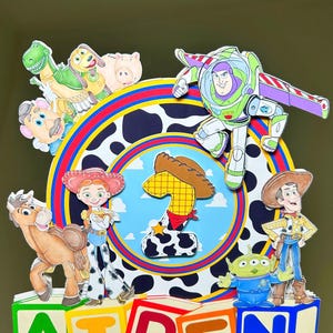 K&ouml;nnte beinhalten: Ein farbenfroher Tortenaufsatz mit Toy Story-Charakteren, darunter Buzz Lightyear und Woody. Der Name "AIDEN" wird in bunten Bl&ouml;cken angezeigt. Das Design beinhaltet einen Kuhdruck und die Zahl eins. Ein rosa Aufkleber mit dem Text "Jams Creations" ist ebenfalls sichtbar.