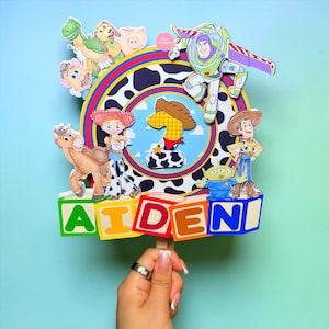 K&ouml;nnte beinhalten: Ein farbenfroher Tortenaufsatz mit Figuren aus dem Film Toy Story. Der Aufsatz zeigt Buzz Lightyear, Woody, Jessie und weitere Charaktere. Der Name "AIDEN" ist in bunten Bl&ouml;cken geschrieben, mit einer Eins in der Mitte.
