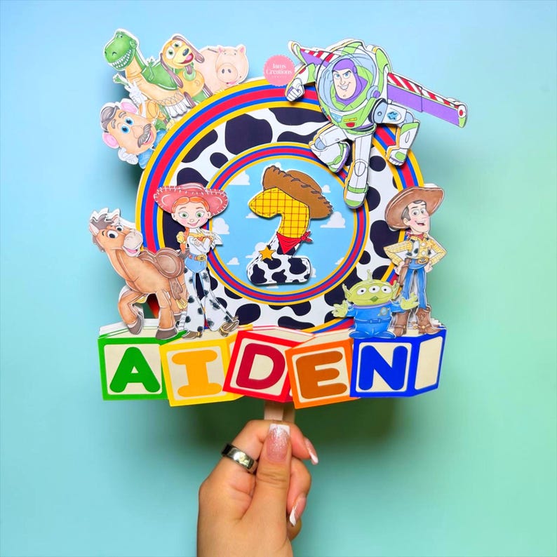 K&ouml;nnte beinhalten: Ein farbenfroher Tortenaufsatz mit Figuren aus dem Film Toy Story. Der Aufsatz zeigt Buzz Lightyear, Woody, Jessie und weitere Charaktere. Der Name "AIDEN" ist in bunten Bl&ouml;cken geschrieben, mit einer Eins in der Mitte.