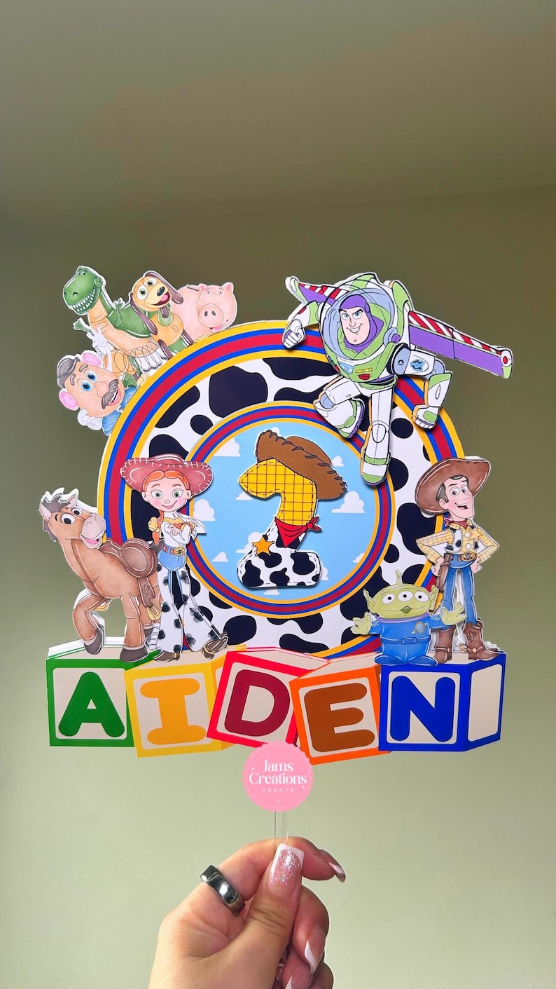K&ouml;nnte beinhalten: Bunte Tortenaufsatz mit Toy Story-Figuren, darunter Woody, Buzz Lightyear und Jessie. Das Design beinhaltet den Namen "AIDEN" in Blockbuchstaben und die Zahl eins. Eine lebendige Dekoration f&uuml;r eine Geburtstagstorte.
