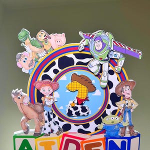 K&ouml;nnte beinhalten: Ein farbenfroher Tortenaufsatz zum Geburtstag mit Toy Story-Charakteren wie Woody, Buzz Lightyear und Jessie. Der Aufsatz zeigt die Zahl eins und den Namen "AIDEN" in Blockbuchstaben. Das Design beinhaltet einen Kuhdruck und einen blauen Hintergrund.