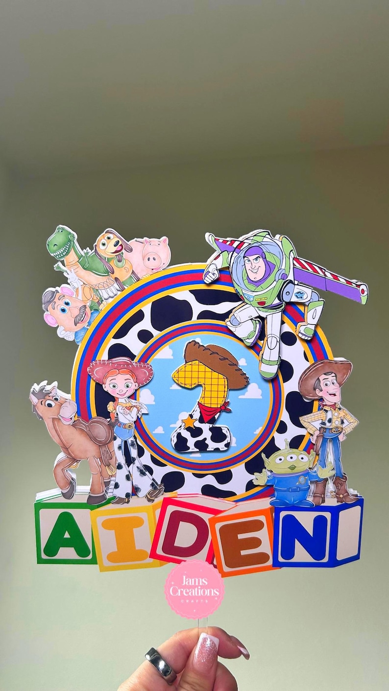 K&ouml;nnte beinhalten: Ein farbenfroher Tortenaufsatz zum Geburtstag mit Toy Story-Charakteren wie Woody, Buzz Lightyear und Jessie. Der Aufsatz zeigt die Zahl eins und den Namen "AIDEN" in Blockbuchstaben. Das Design beinhaltet einen Kuhdruck und einen blauen Hintergrund.