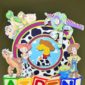 K&ouml;nnte beinhalten: Bunte Tortenaufsatz mit Toy Story-Figuren, darunter Buzz Lightyear und Woody. Das Design enth&auml;lt den Namen "AIDEN" in bunten Bl&ouml;cken. Ein rosa Aufkleber mit dem Text "Jams Creations" ist ebenfalls sichtbar. Thema: Geburtstag.