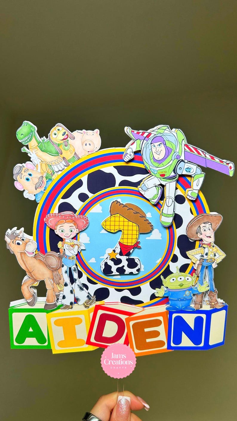 K&ouml;nnte beinhalten: Bunte Tortenaufsatz mit Toy Story-Figuren, darunter Buzz Lightyear und Woody. Das Design enth&auml;lt den Namen "AIDEN" in bunten Bl&ouml;cken. Ein rosa Aufkleber mit dem Text "Jams Creations" ist ebenfalls sichtbar. Thema: Geburtstag.