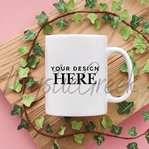Puede incluir: Una taza de cerámica blanca con asa, con el texto "YOUR DESIGN HERE" en negro. La taza está colocada sobre una tabla de madera, rodeada de una vid decorativa con hojas verdes. El fondo es rosa suave.