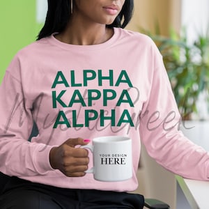 Puede incluir: Una sudadera rosa claro con el texto "ALPHA KAPPA ALPHA" en verde. Se sostiene una taza blanca con el texto "YOUR DESIGN HERE". La sudadera tiene mangas largas.