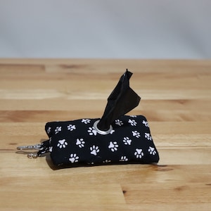 May include: A black dog poo bag dispenser with a white paw print pattern. The dispenser has a silver ring and a black plastic bag is partially pulled out.