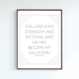 May include: A framed print with a white background and the text "THE LORD IS MY STRENGTH AND MY SONG, AND HE HAS BECOME MY SALVATION" in black. The frame is a muted gray, and the text is accented with a pale pink geometric design.