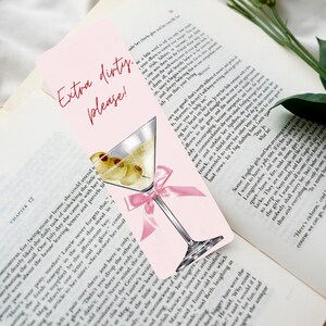 May include: Pink bookmark with a martini glass illustration, olives, and a pink bow. Text reads "Extra dirty, please!". The bookmark is set against the backdrop of an open book.
