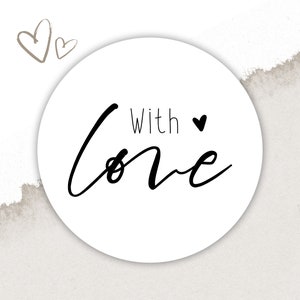 以下が含まれることがあります： 白い円の中に、黒い文字で「With love」と小さな黒いハートが書かれています。背景は薄い茶色のテクスチャのある紙で、左上の隅に小さな茶色のハートが2つあります。