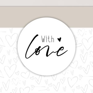 以下が含まれることがあります： 黒い筆記体で「With Love」と書かれた白い丸いステッカーと小さな黒いハート