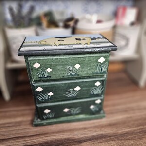 May include: A small, green wooden chest of drawers with three levels. The chest is decorated with white flower and grass designs. The top of the chest features a light blue background with a yellow animal illustration.