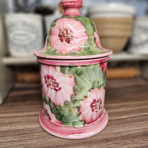 Può includere: Un vaso in ceramica rosa e verde con coperchio, con un design floreale. Il vaso ha un corpo cilindrico e una base svasata. Il coperchio è a forma di campana con un piccolo pomello. Il motivo floreale comprende fiori rosa e foglie verdi.