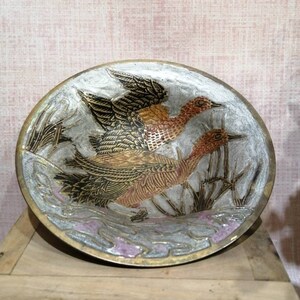 Puede incluir: Un plato redondo de latón con un fondo plateado y un diseño en relieve de dos pájaros en vuelo. Los pájaros están pintados en tonos de marrón, dorado y negro, y el fondo está decorado con un patrón floral estilizado.