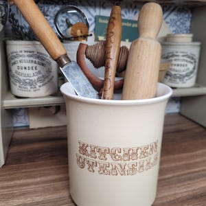 May include: A white ceramic kitchen utensil holder with the words "KITCHEN UTENSILS" printed in brown. The holder contains a wooden rolling pin, a wooden spoon, a wooden dowel, and a silver butter knife.
