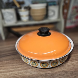 May include: A round enamel casserole dish with a bright orange lid featuring a black knob. The base of the dish is white and decorated with a repeating pattern of orange, yellow, and green circular floral motifs with a thin silver rim.