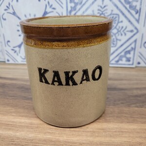 May include: A ceramic jar with a brown rim and a white body. The jar has the word "KAKAO" written in black lettering.