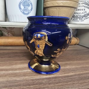 May include: A dark blue ceramic vase with gold accents. The vase features a pedestal base and a rounded body with a detailed gold figure and floral designs. The vase is approximately 6 inches tall.