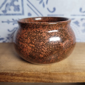 May include: A small, round, brown ceramic pot with a speckled orange glaze. The pot is sitting on a wooden surface.