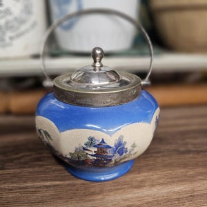 May include: A small, blue and white ceramic sugar bowl with a silver lid and handle. The bowl features a landscape design with trees and buildings. The lid has a small, round knob. The bowl is sitting on a wooden surface.