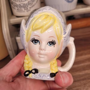 May include: A ceramic mug shaped like a girl's head with blonde braids, blue eyes, and a blue and white dress. The mug has a handle on the side.