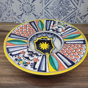 Op de afbeelding: Een ronde keramische schaal met een kleurrijk ontwerp. De schaal heeft een gele rand, blauwe en witte strepen en een centraal ontwerp met een zon, bladeren en een schild. De schaal is versierd met rode, oranje en groene accenten.