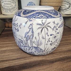 Op de afbeelding: Een ronde, witte keramische vaas met een blauw en wit pastorale scène. De vaas toont een hert, palmbomen en bloemmotieven. De rand en de bovenkant zijn ook versierd met blauwe patronen. De vaas staat op een houten oppervlak.