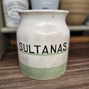 Può includere: Un vaso di ceramica bianco con la scritta "SULTANAS" in nero. Il vaso presenta una fascia a righe verdi al centro e poggia su una superficie di legno. Altri contenitori in ceramica sono visibili sullo sfondo.