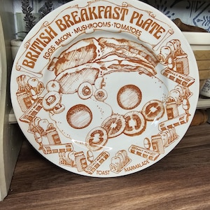 Op de afbeelding: Een witte keramische plaat met een bruine illustratie van een Engels ontbijt. De plaat toont eieren, spek, champignons en tomaten. De tekst op de plaat luidt "British Breakfast Plate" en bevat de woorden "Eggs, Bacon, Mushrooms, Tomatoes, Toast, Marmalade".