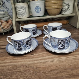 May include: Four matching teacups and saucers. The cups and saucers are white with a blue and silver design. The design includes a celestial pattern. The cups have a curved handle. The saucers are round with a silver trim.