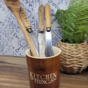 Op de afbeelding: Een bruine keramische bestekhouder met de tekst "Kitchen Things" aan de voorkant. De houder bevat een houten lepel en drie messen met houten handvatten.