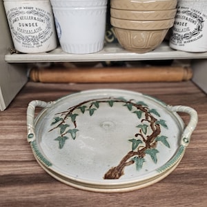 Peut inclure: Un plateau de service rond en céramique avec des poignées, orné d'un motif de lierre peint à la main en vert et marron. Le plateau a un fond bleu-vert clair et une bordure décorative. Le plateau est sur une surface en bois.