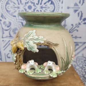 May include: A ceramic frog-themed candle holder with a green glaze and a cut-out design. Two frogs are perched on the edge of the holder, which is decorated with leaves and vines.